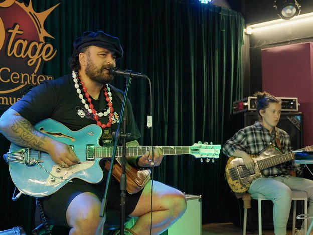 A man sits on a chair on a stage playing a guitar and singing into a microphone in a music venue. A female performer sits behind him playing guitar.