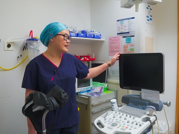 A female doctor stands in a hospital room pointing at some medical equipment. She wears a blue hairnet.