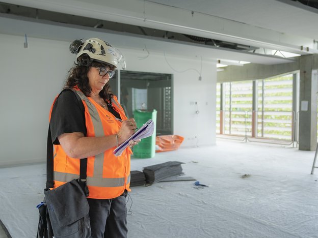 A woman writes notes while she wears a hardhat and high visibility vest.