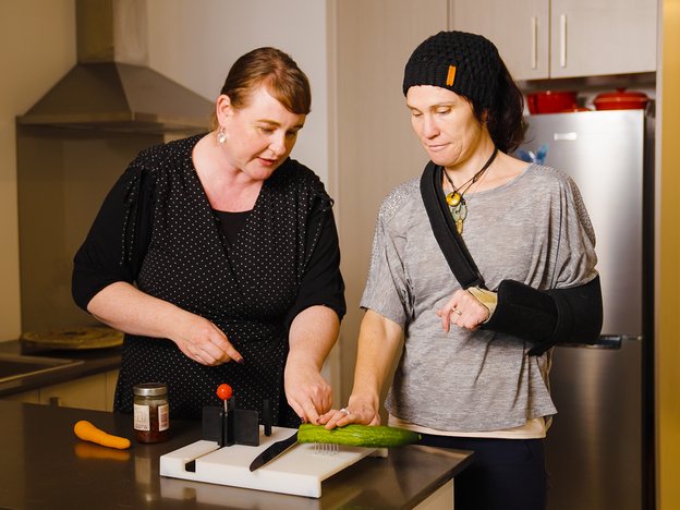 A woman shows another person how to chop a cucumber using an adaptive cutting board.
