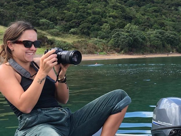 A female photographer uses a camera to take a photo. She sits on a boat in a river.