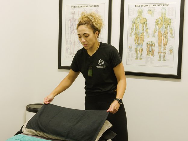 A woman places a black towel onto a pillow on a physiotherapy bed. There are two pictures of the human body on the wall behind her.
