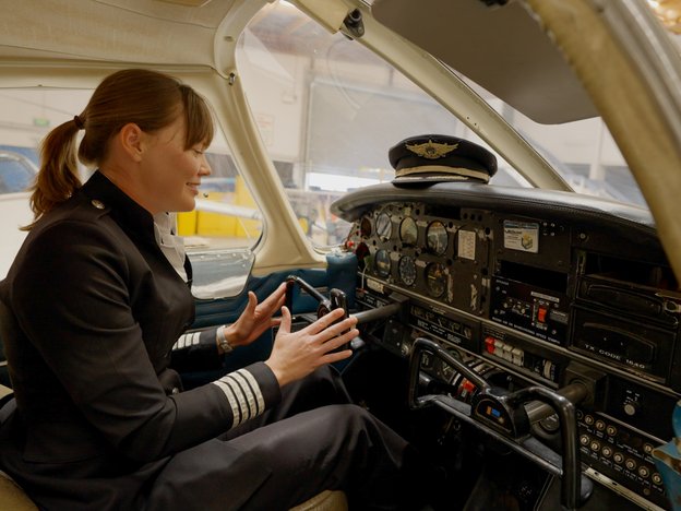 A female pilot sits in the front seat of an airplane. She holds the steering wheel and is wearing a pilot uniform. There is a pilot cap on the dashboard.
