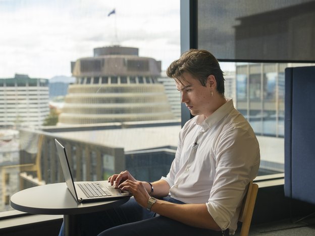 A man sits on a chair at a small, round table in front of a large, glass window. He uses a laptop. The beehive is in the background view.