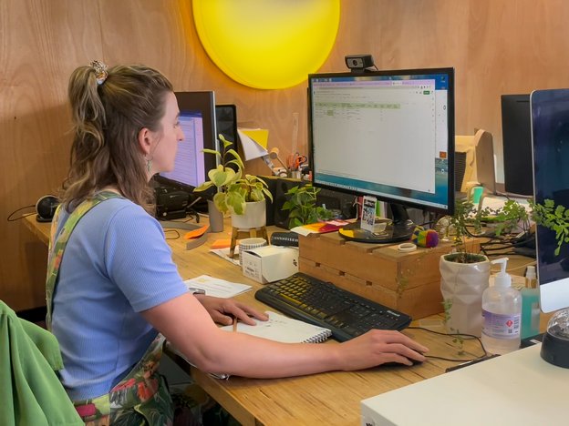 A woman in an office wears a blue shirt and colourful dress. She sits at a wooden desk in front of a monitor.