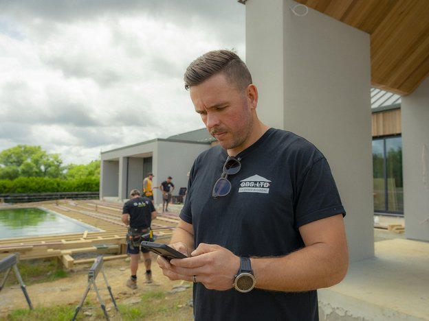 A man stands at a construction site. He uses a mobile phone. There are other construction workers behind him.