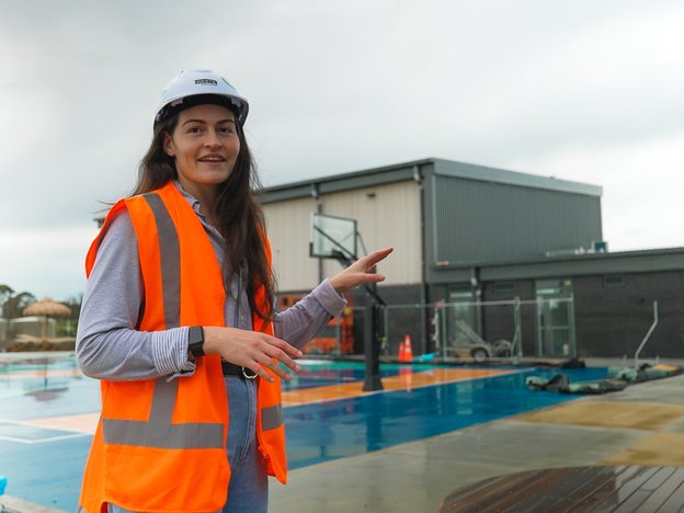 A woman stands outside on a basketball court looking into the camera. She wears an orange vest and white hard hat.