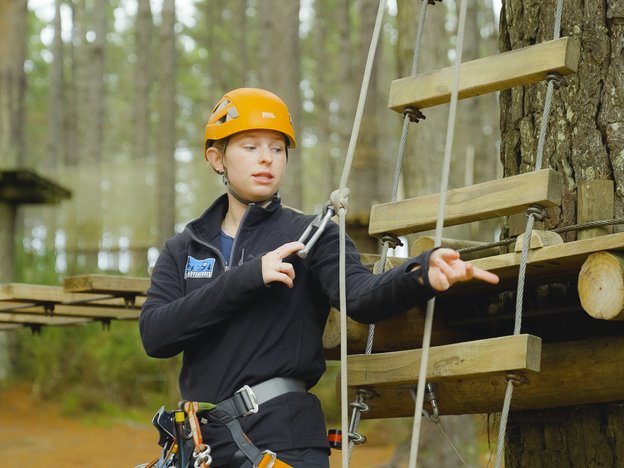 An outdoor recreation professional instructs how to use a ladder and ropes course. They are wearing an orange safety helmet.