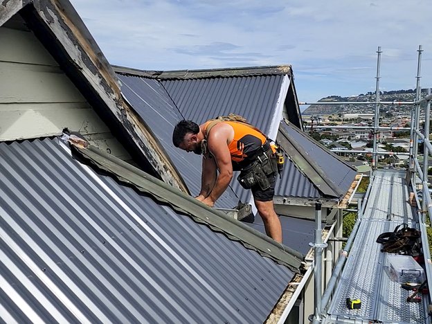 A roofer repairs a roof. They are wearing an orange hi-vis singlet, shorts and a toolbelt, next to the roof is scaffolding.