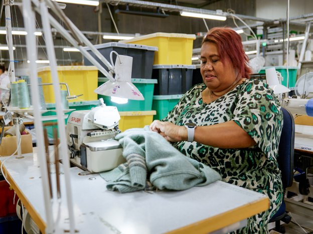 A sewing machinist is sitting at an overlocker holding a woollen garment.