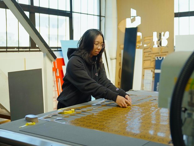 A woman stands in a workshop working on a large, brown sheet of signage laid out on a table in front of her.
