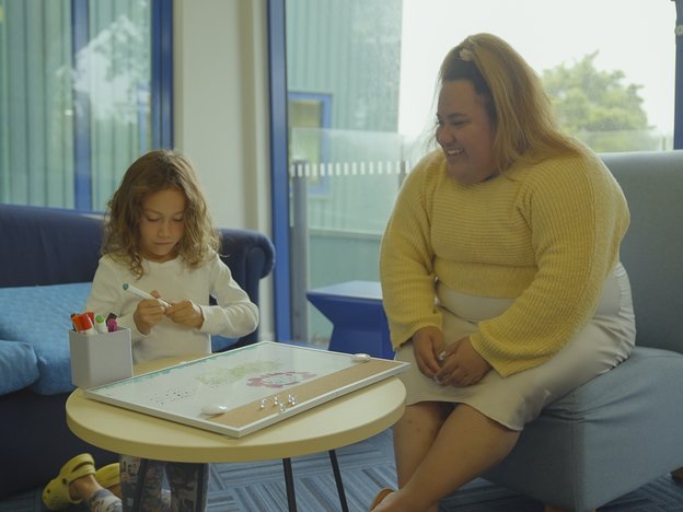 A female social worker sits on a chair in a meeting room. She is with a young girl kneeling on the ground in front of a table. They use colourful pens on a whiteboard.