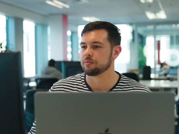 A young man looks at a laptop and computer monitor screen in front of him. He is in an office.