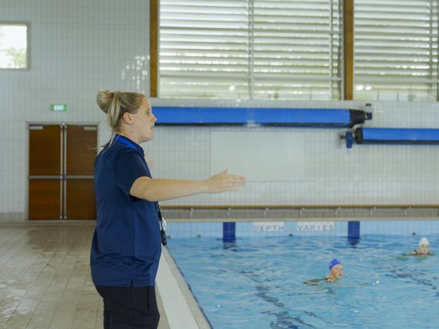 A woman stands with raised arms stands next to a large blue pool coaching two young people in a swimming pool. She wears a navy uniform.