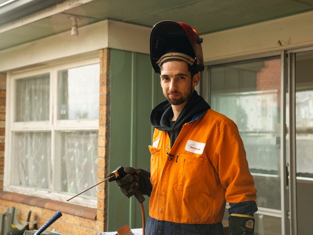 A man stands outside a house holding a fabrication tool. He wears orange overalls and a protective face shield.