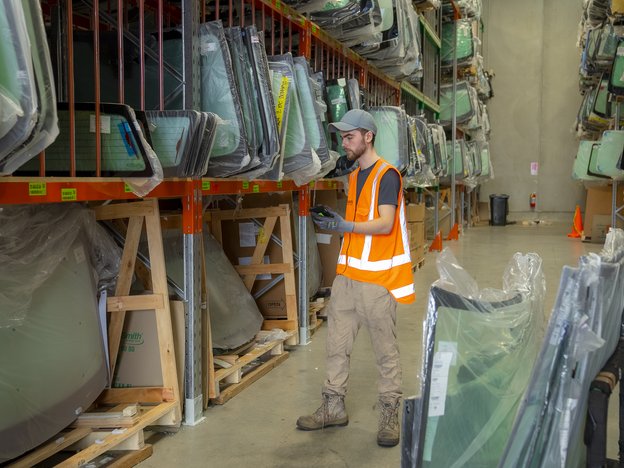 A man stands in in the backroom of a windscreen factory. He wears an orange vest, cap and work boots.