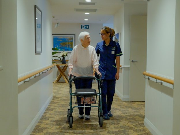 A medical worker helps an elderly person walk down a hallway. The elderly person is also using a stroller.