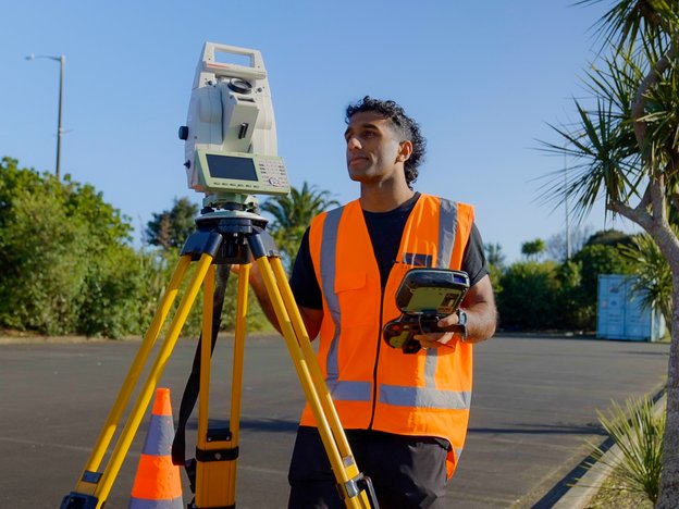 A surveyor observes their tripod. They are in a sunny car park.