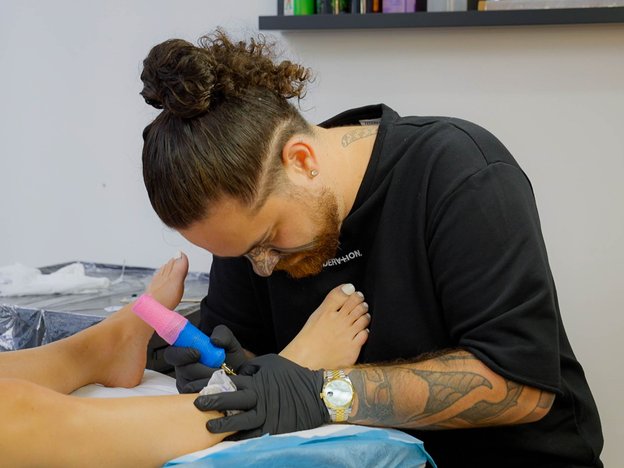 A male leans over a person's feet holding a tattoo gun. He tattoos their foot. He wears a black t-shirt and gloves and has a ponytail.