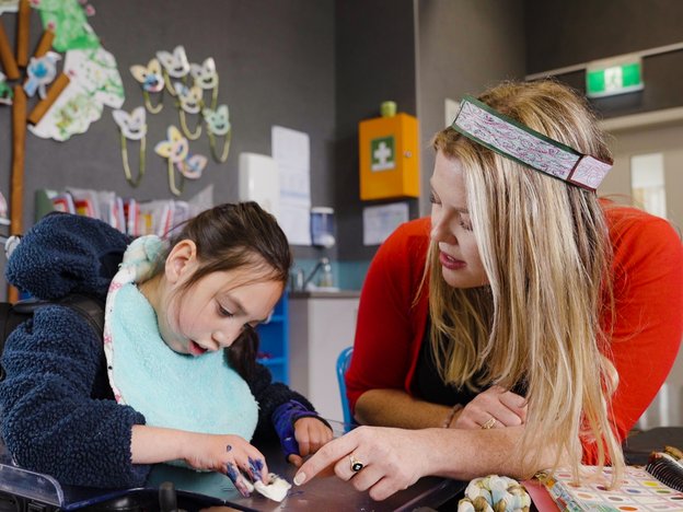 A woman helps a young girl in a wheelchair. They sit at a table. The woman points to something the girl is playing with.