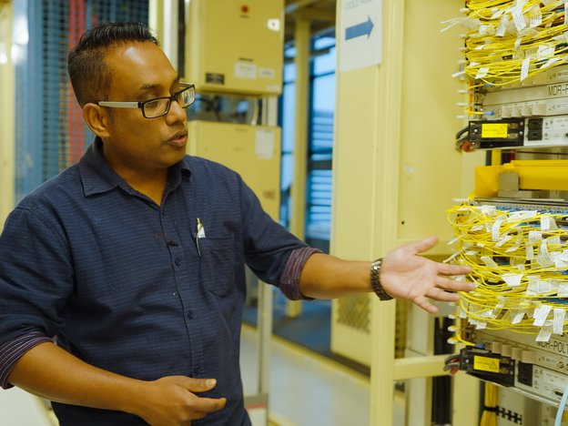 A man points to yellow wires sticking out of a wall in a factory. He wears glasses.