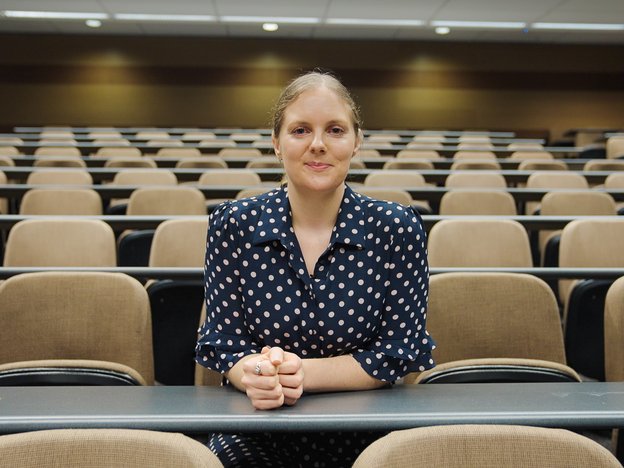 A smiling woman sits on a chair in a row of seats in a lecture hall.