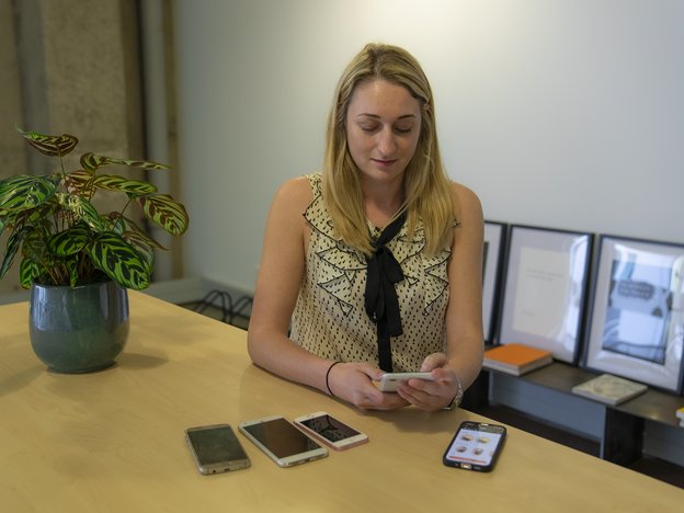 A woman sits at a table holding a mobile phone. There are four other phones in front of her. She is in an office.