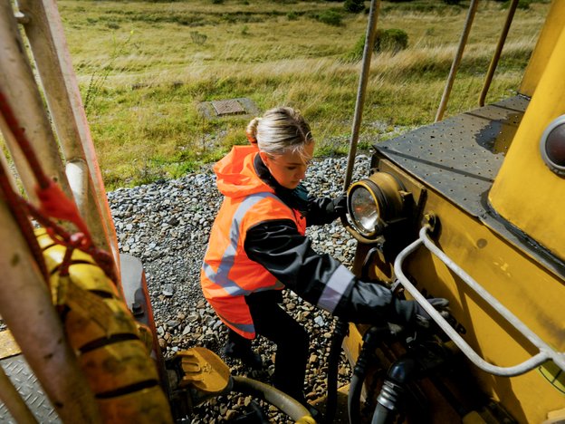 A female train manager wearing hi-vis and working outside between two carriages. There is grassland in the background.