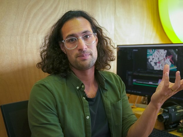 A man with glasses gestures in a trendy office space. He edits a video on a computer.