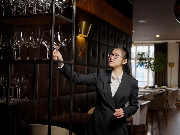 A female waitress stands in a restaurant. She holds a wine glass from a wine shelf. She wears a black suit and white shirt.