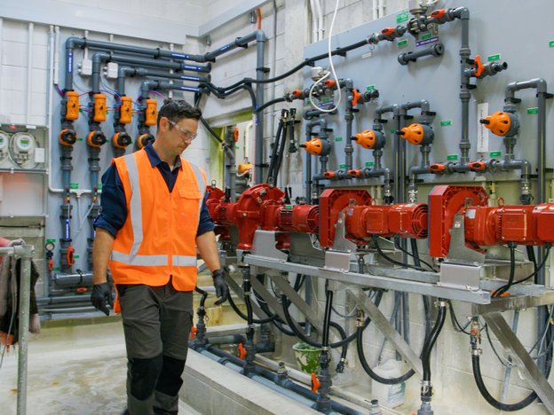 A water treatment specialist wearing a hi-vis vest and safety glasses is looking at a complex system of pipes in the treatment plant.