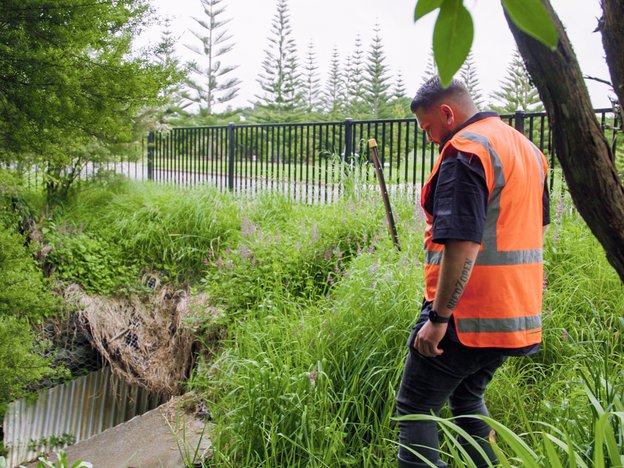 A water resource specialist wearing hi-vis vest is amongst grass looking at a culvert beside the road.