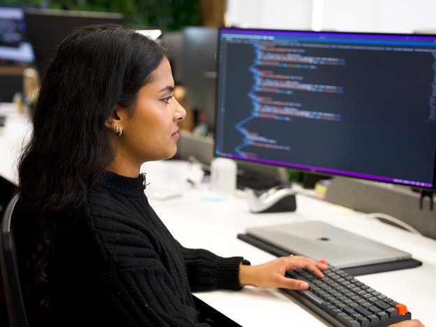 A young web developer sits in office focused on her computer. Code can be seen on one of her monitors.