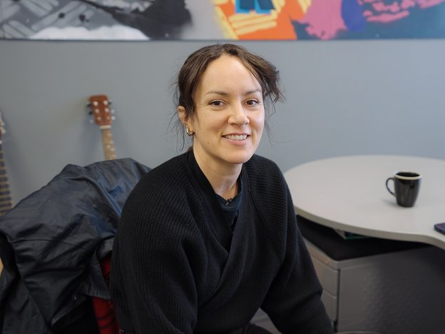 A woman sits in a chair in an office smiling into the camera. There are two guitars behind her.