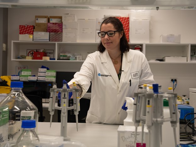 A scientist takes a pipette from an auto pipette stand. They are wearing a white lab coat and glasses.
