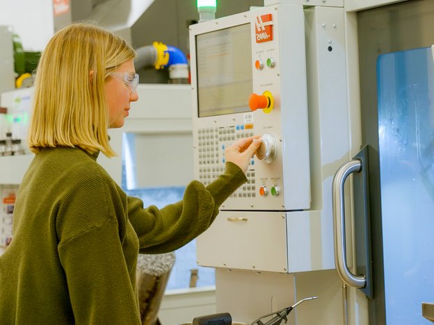 Woman with blonde hair wears an olive green jersey and protective glasses. She reaches for a dial on a piece of scientific equipment.