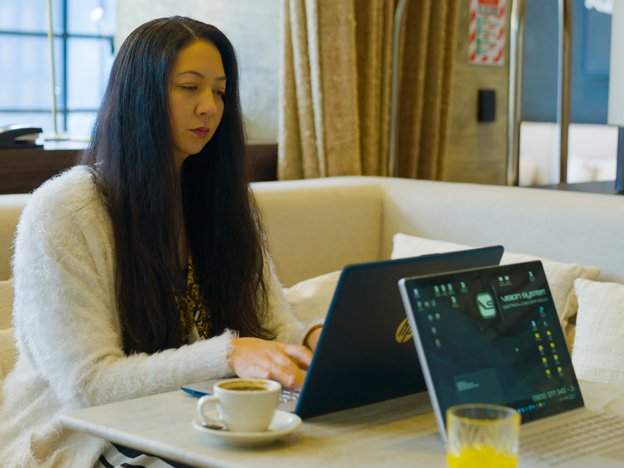 A woman sits at a table typing on a laptop in an office. There is a cup of coffee next to her, and another person's laptop in front of her.
