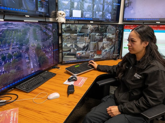 A woman sits at a desk monitoring video footage on six computer screens.