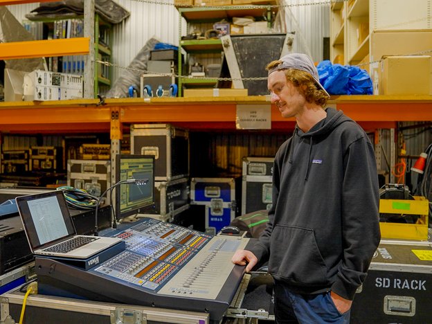 A technician uses audio control equipment. They are surrounded by boxes of stage equipment.