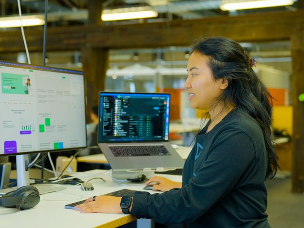 A young woman sits at a desk looking at a computer monitor and laptop. She is using a mouse and keyboard.