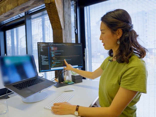 A young woman sits at a desk using a laptop, mouse and keyboard. She points to some data on a computer screen in front of her.