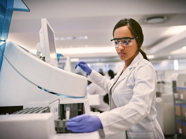 A female scientist uses a machine in a laboratory. She is wearing a white lab coat, blue gloves and safety glasses.