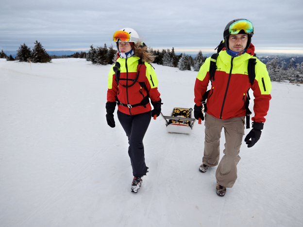 Two ski patrollers carry a first aid sled up a ski slope.