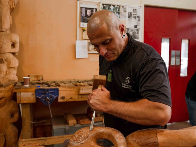 E tū ana he tāne i roto i tētahi taiwhanga mahi e whakairo ana i tētahi wāhanga rākau hei waihanga i tētahi mahi toi Māori. E whakamahi ana i te hama me te whao.