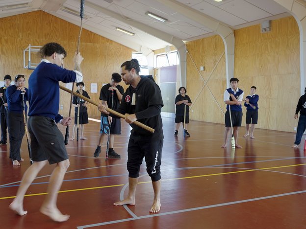E tohutohu ana he tāne i te mau taiaha ki tētahi rōpū ākonga tāne kura tuarua. E whakawai ana rātou i te hōro kura.