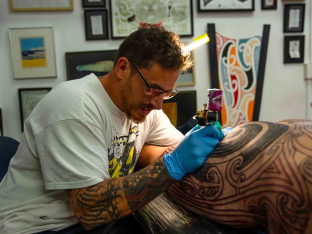 E noho ana tētahi tāne i roto i tētahi taiwhanga tāmoko e tā ana i te moko ki te tinana o tētahi tangata. E pupuri ana ia i tētahi pūrere tāmoko, e mau komoringa kahurangi ana hoki.