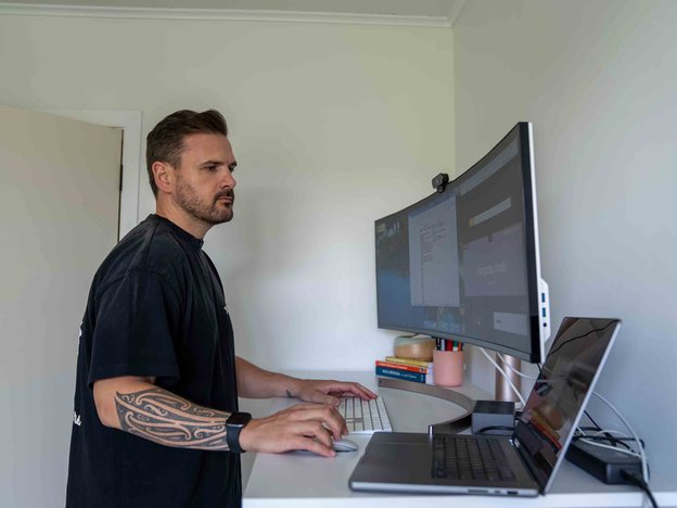 A man stands at his desk in his home office. He has two monitors, a laptop and mouse in front of him.