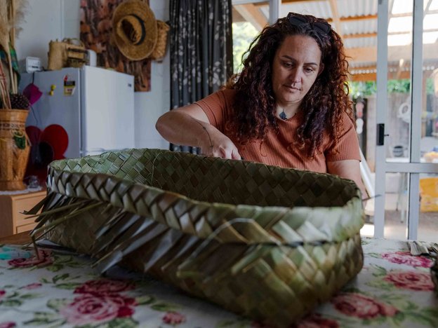 E tū ana he wahine i te taha o tētahi tēpu i tētahi wāhi noho e raranga ana i tētahi kete ki ana matimati.