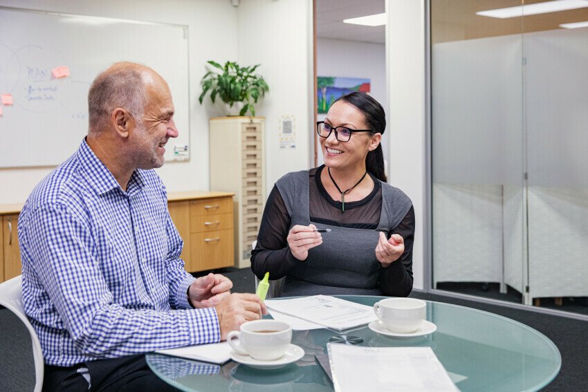 A man and a woman sit in an office at a small, glass, round table with coffee mugs and papers on it.