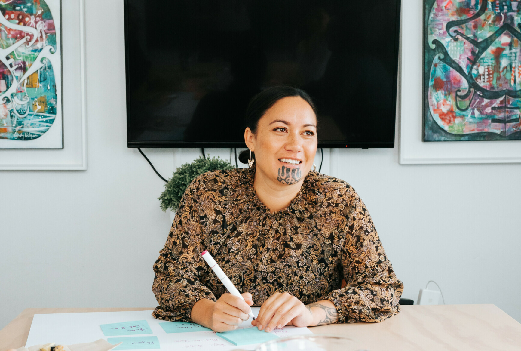 A person sits at a work table. They are holding a pen are about to write on a post-it note.